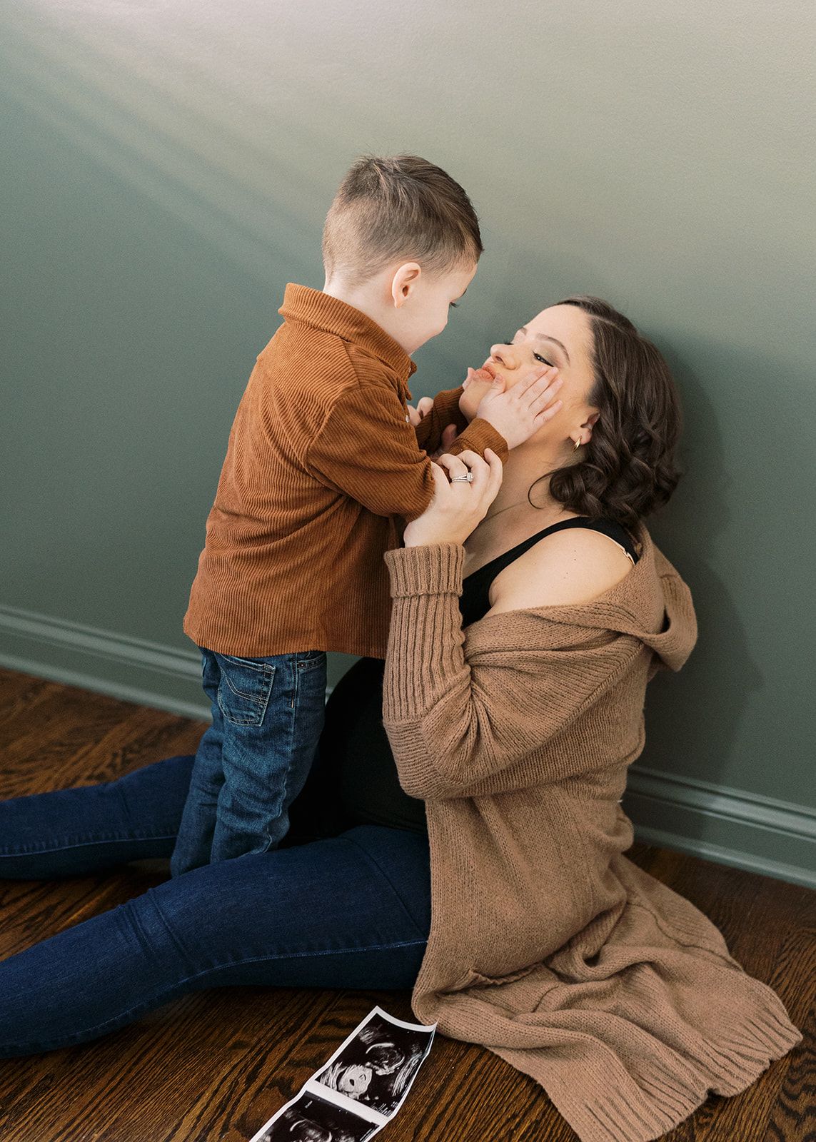 A mother kneels on a hardwood floor smiling at her young son, with ultrasound photos displayed in front of them.