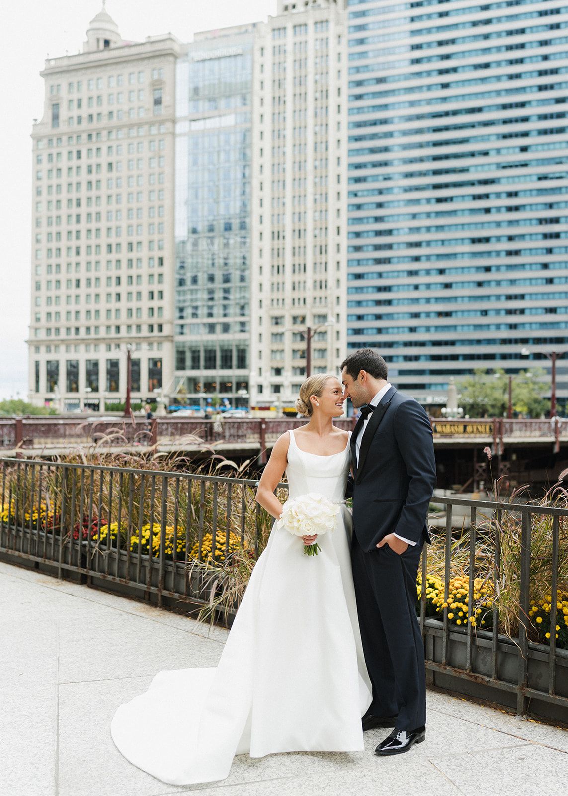 Wedding couple posing on Chicago riverwalk with city skyline backdrop, bride in white gown holding bouquet.