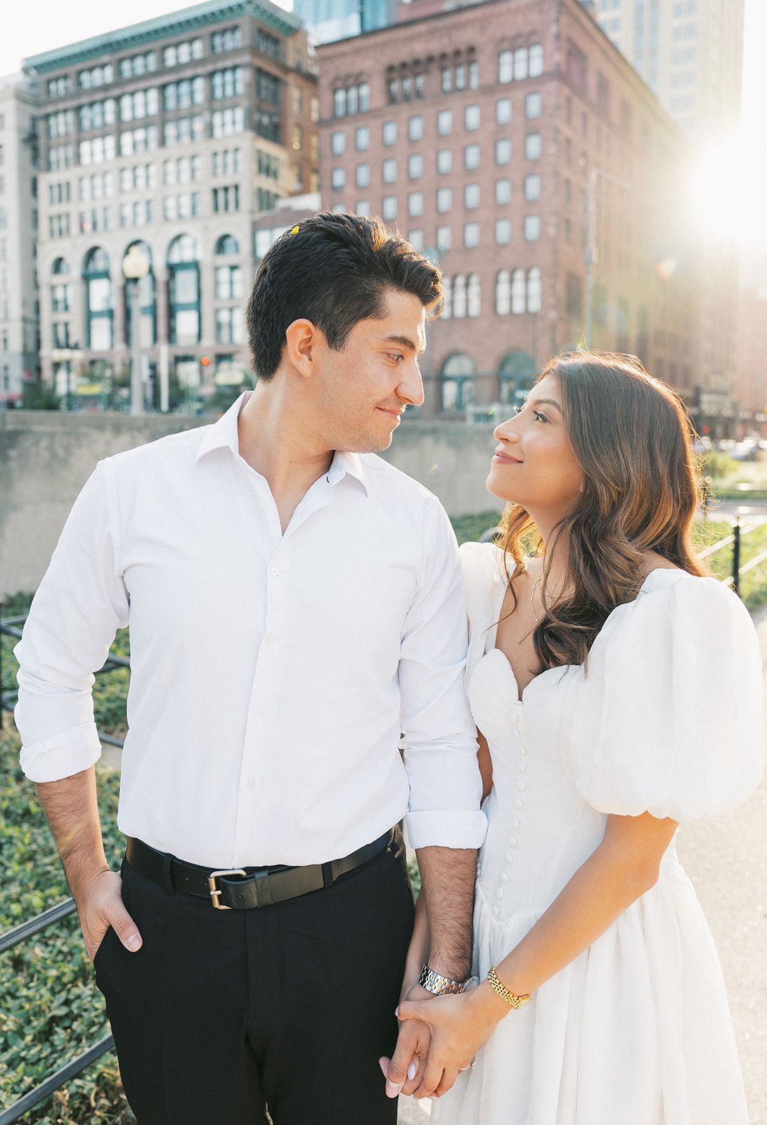 Couple in white outfits holding hands and gazing at each other with sunlit brick buildings in the background.