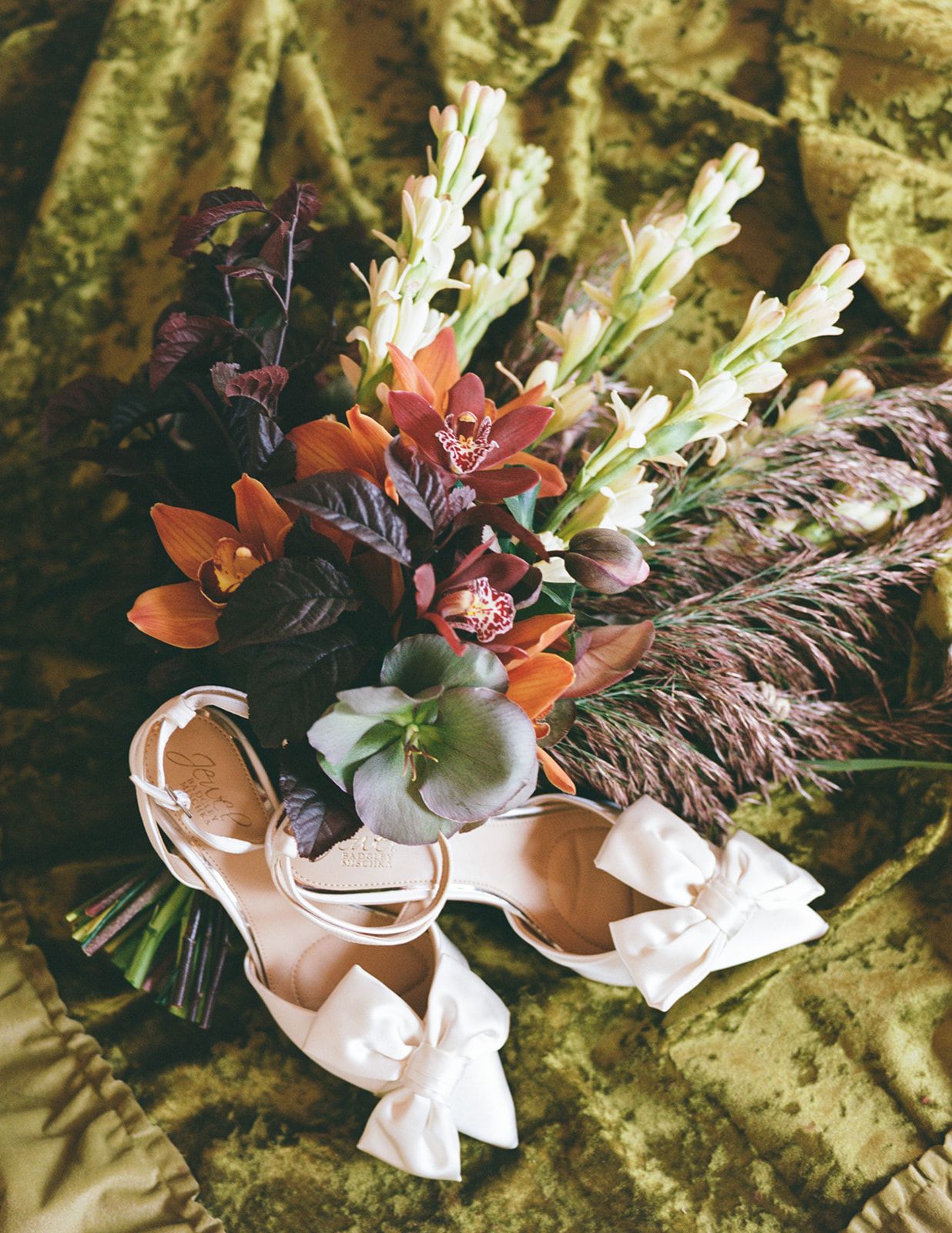 Bridal bouquet with orange orchids and wildflowers beside white bow heels on green velvet fabric.