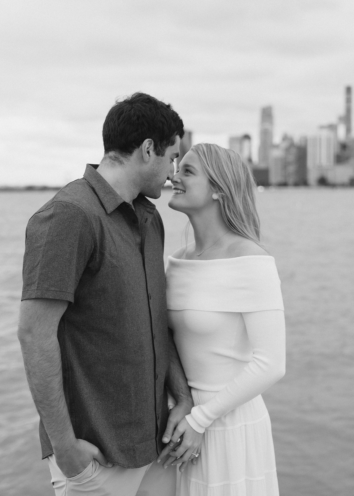 Black and white photo of a couple smiling at each other near a city waterfront.