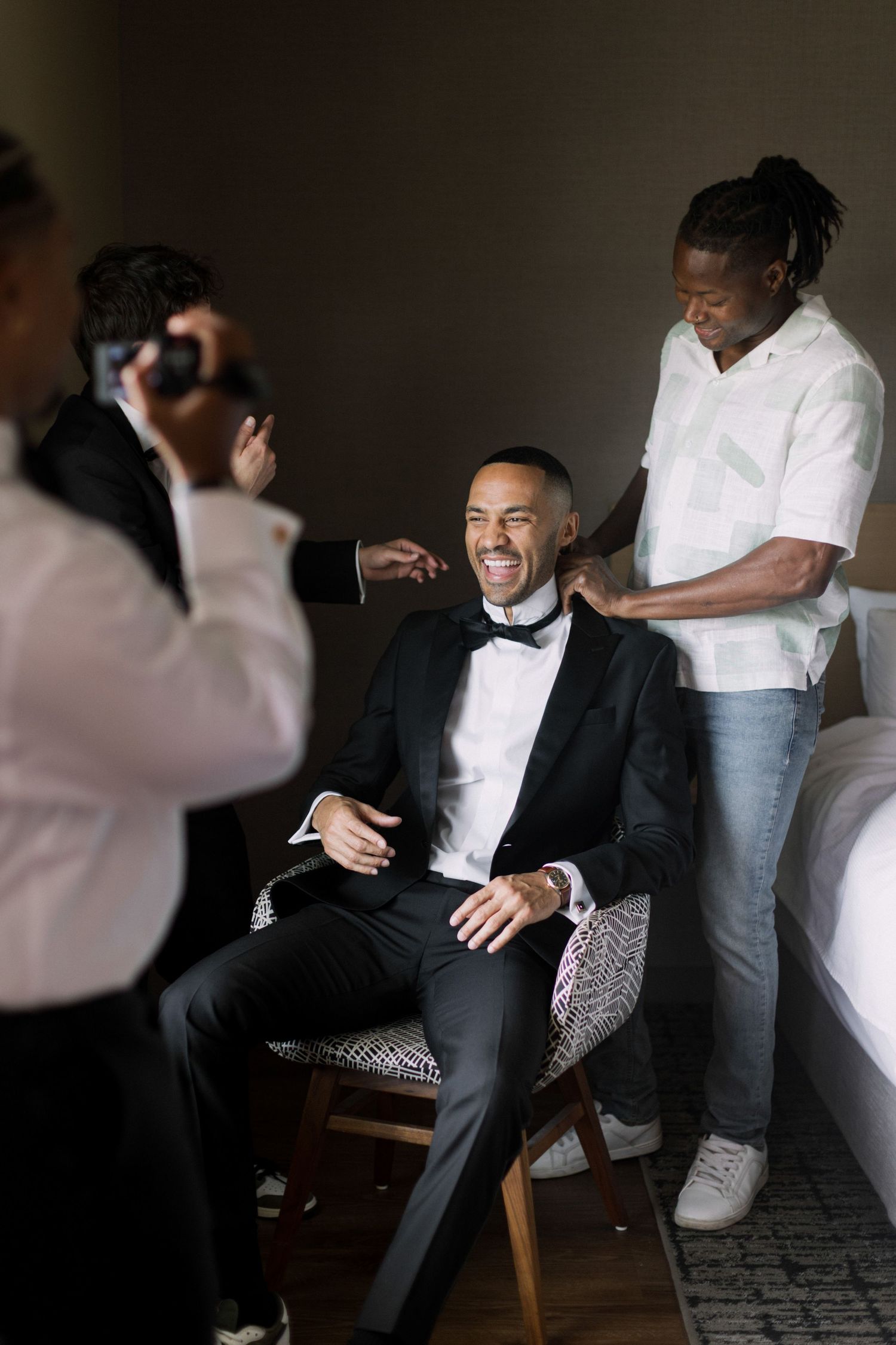 Groom in tuxedo laughing while getting ready with groomsmen, photographer capturing candid wedding preparation moment.