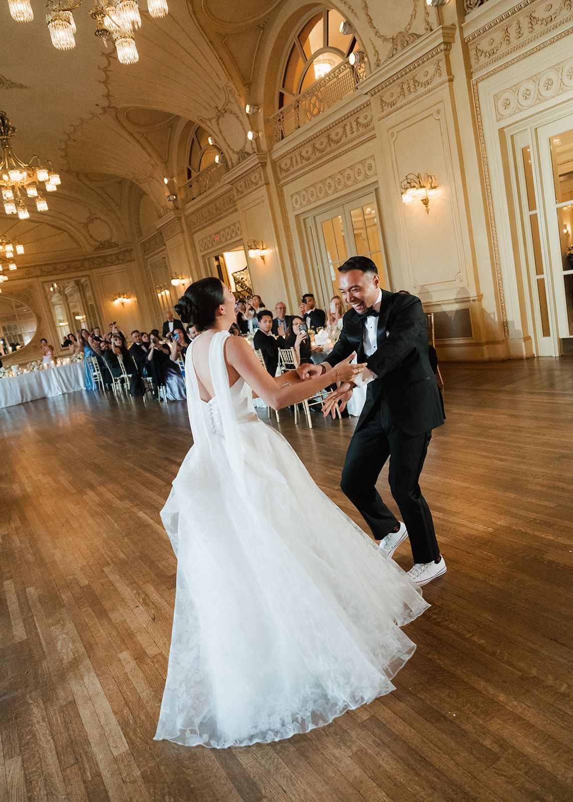 Bride and groom share their first dance in an elegant ballroom with ornate gold decor and guests watching.