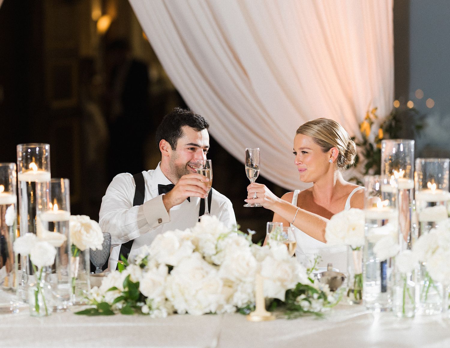 Bride and groom toasting with champagne at sweetheart table adorned with white floral centerpieces.