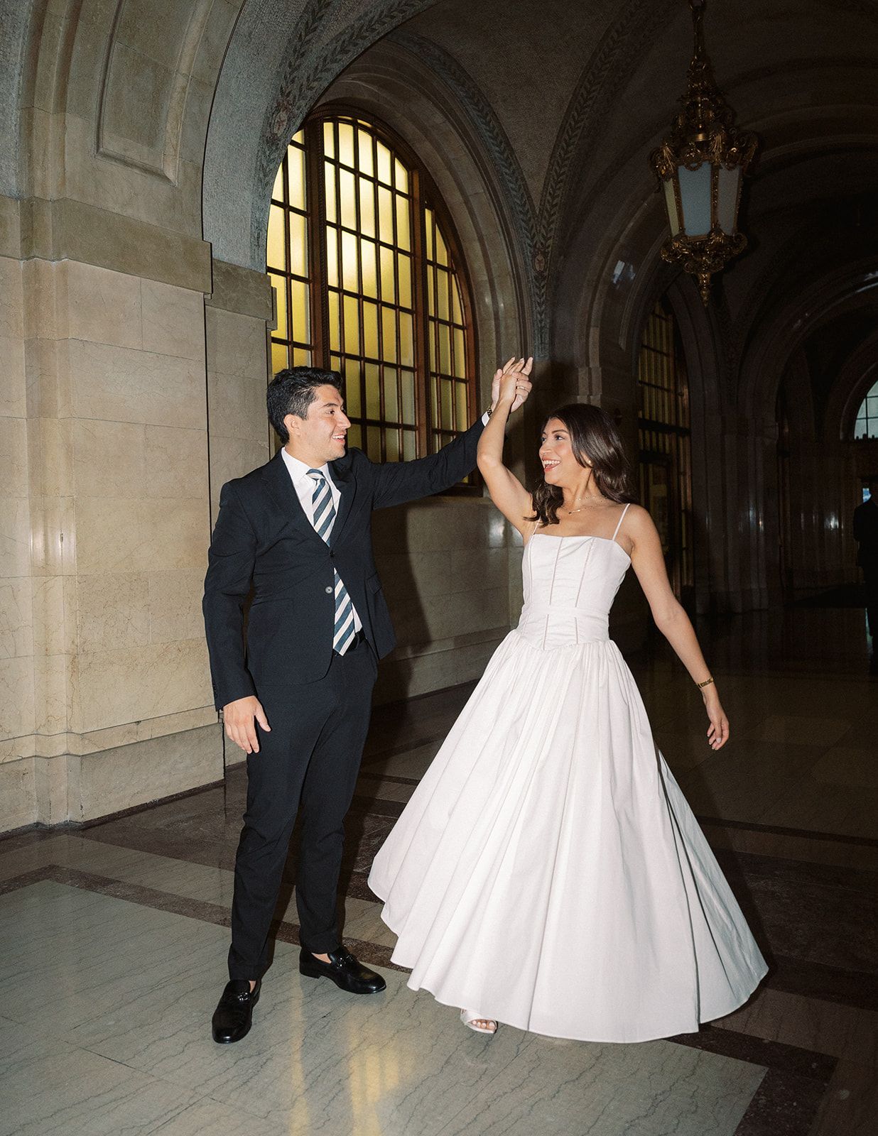 Bride in white gown and groom in navy suit share a joyful high-five in an elegant arched hallway.