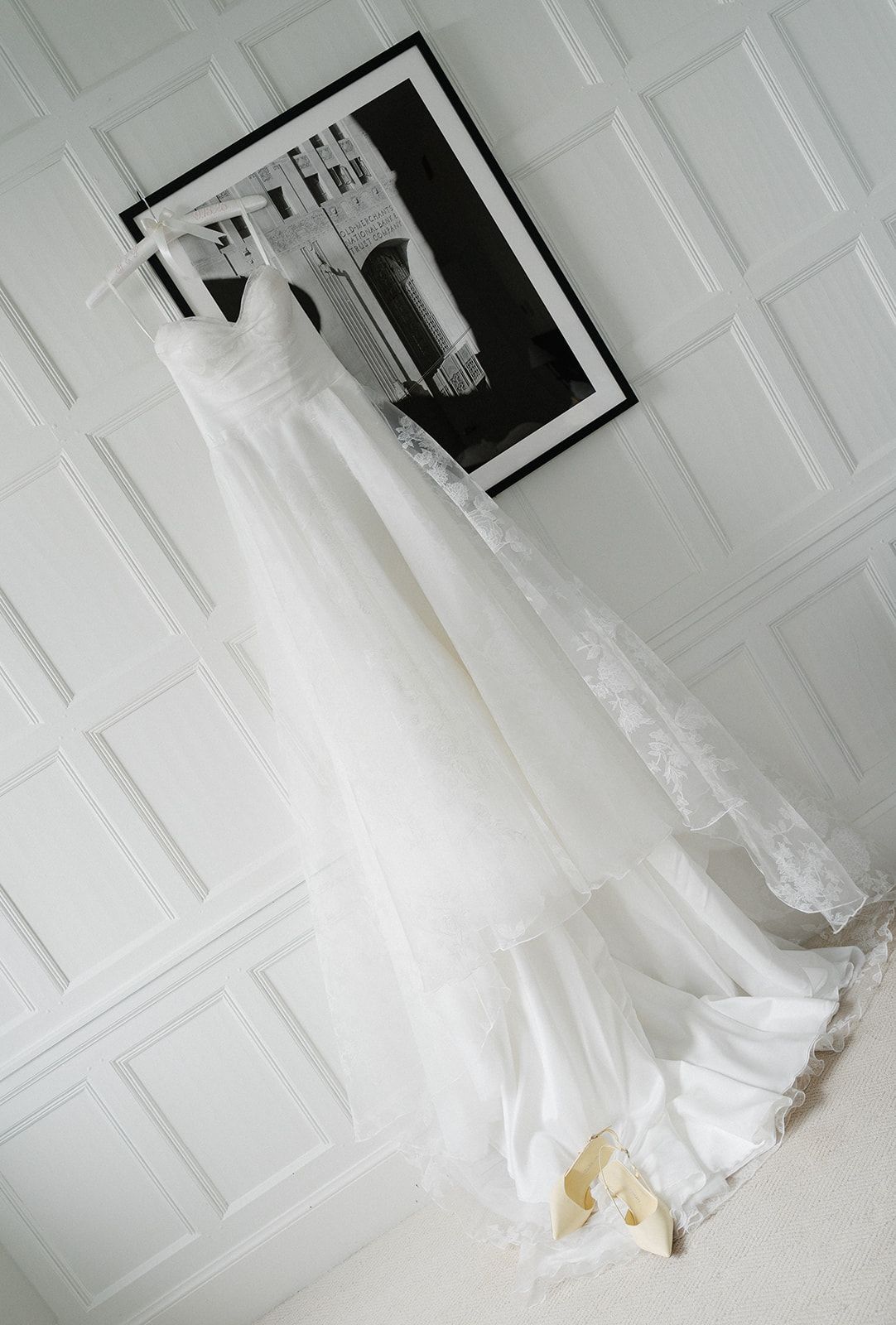 White wedding dress and veil hanging elegantly against a paneled wall with bridal shoes displayed below.