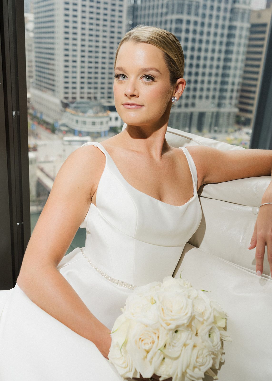 Beautiful blonde bride in sleek white gown holds white rose bouquet beside city skyline window.