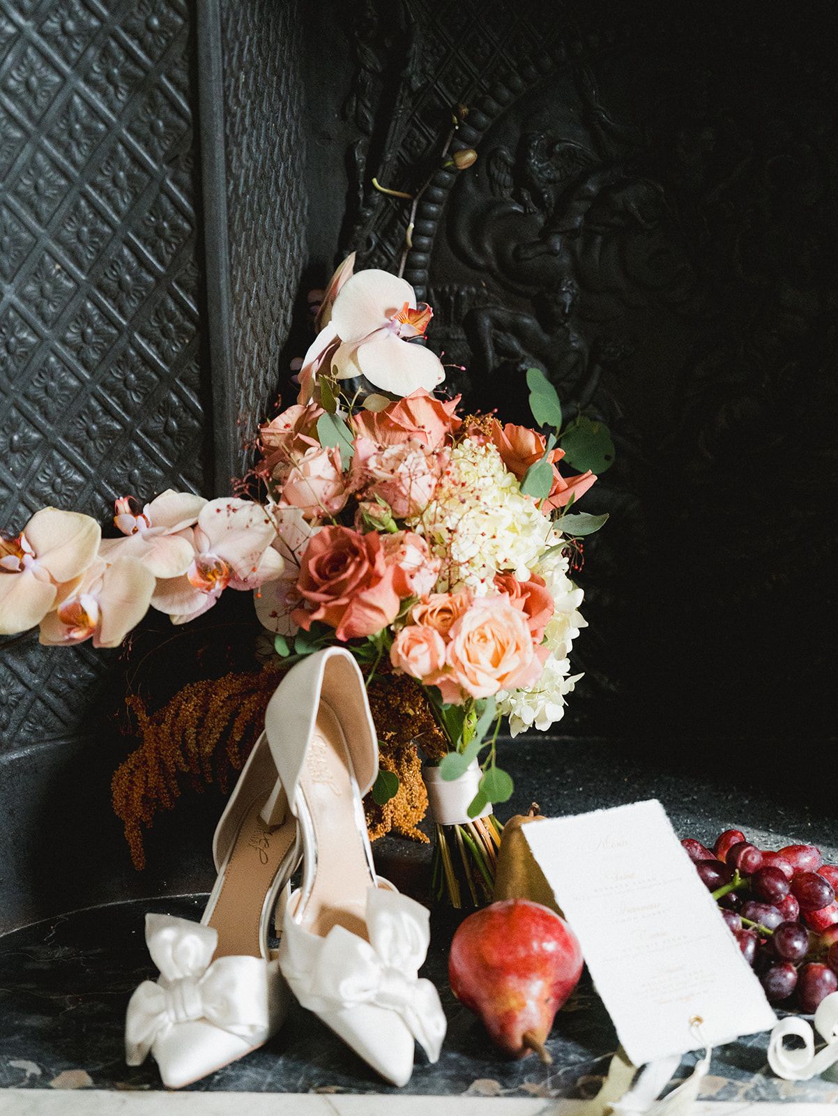 White bow heels beside peach roses, orchids, pomegranate, grapes, and wedding stationery on dark surface.