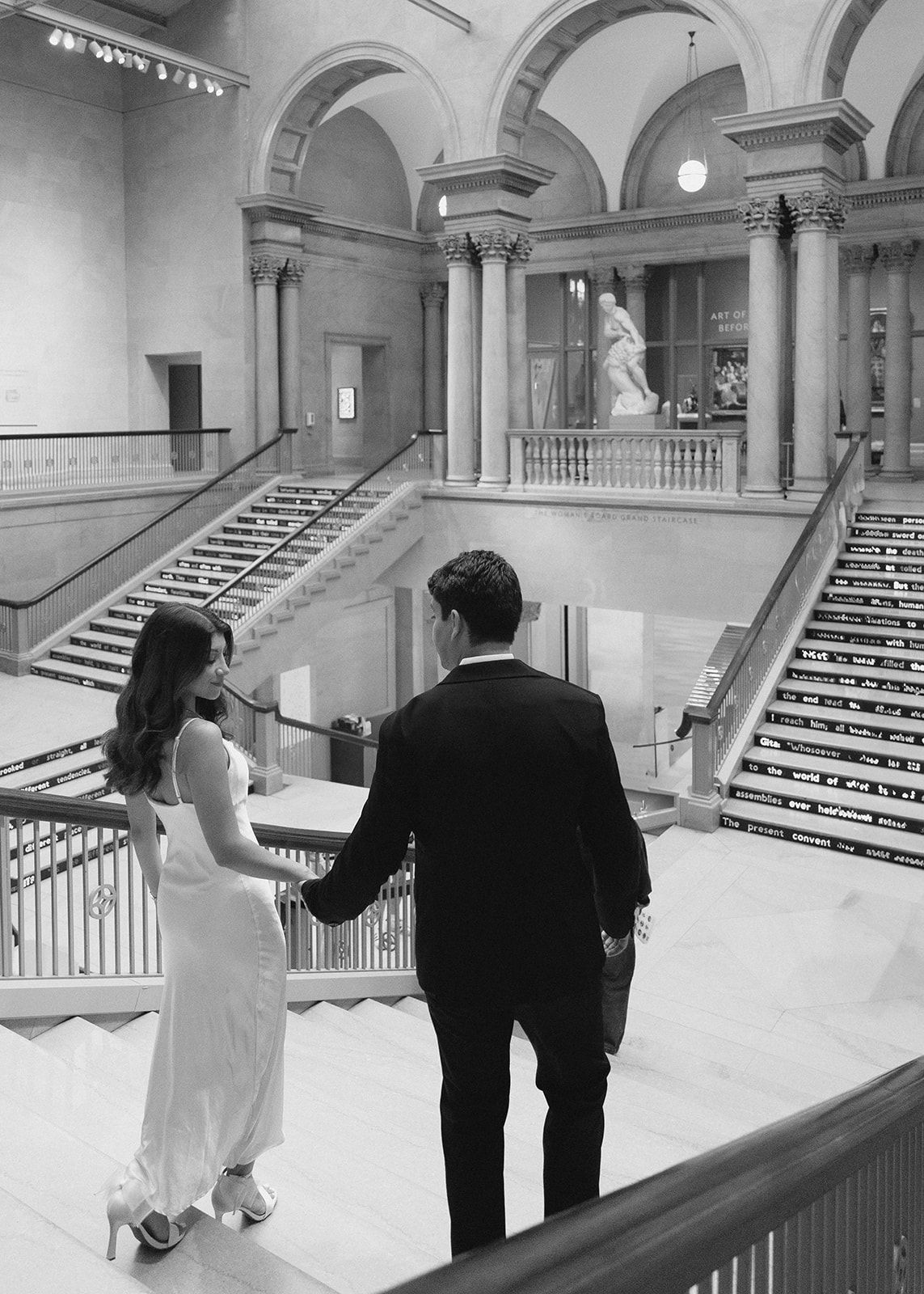 Couple holding hands in elegant museum interior with grand staircase and classical statue in black and white.