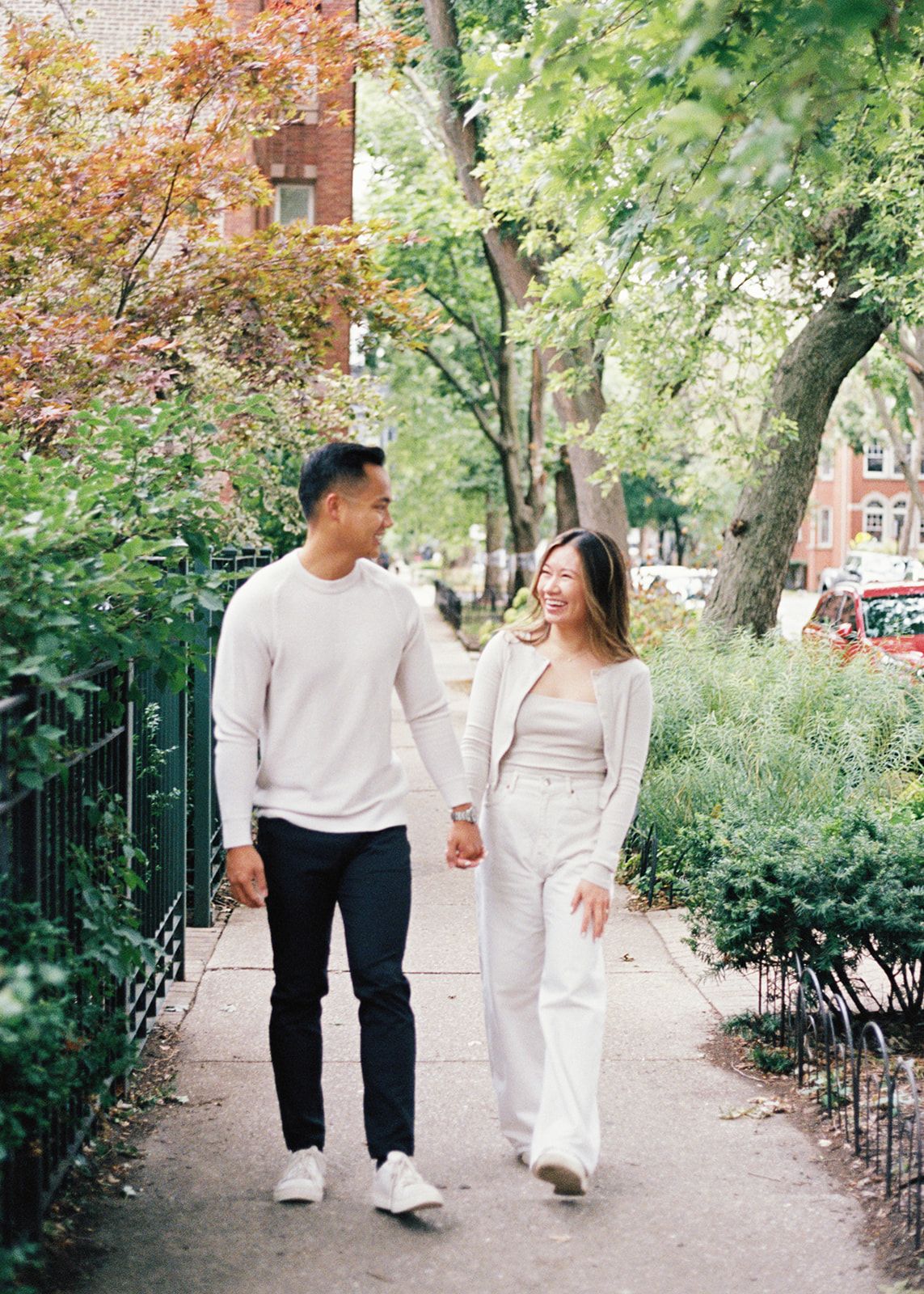 Happy Asian couple walking hand in hand on tree-lined urban sidewalk in neutral outfits.