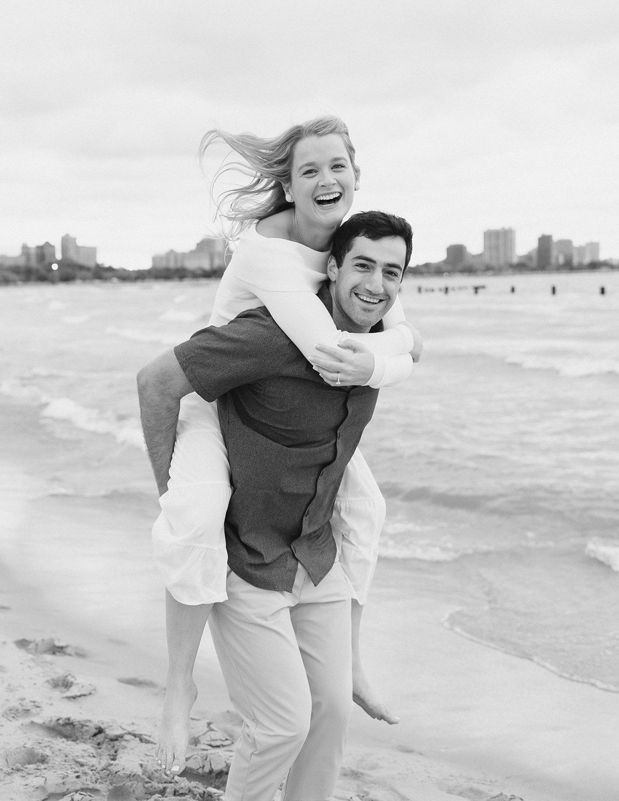 Joyful couple laughing on a beach as man gives woman a piggyback ride near the ocean waves.