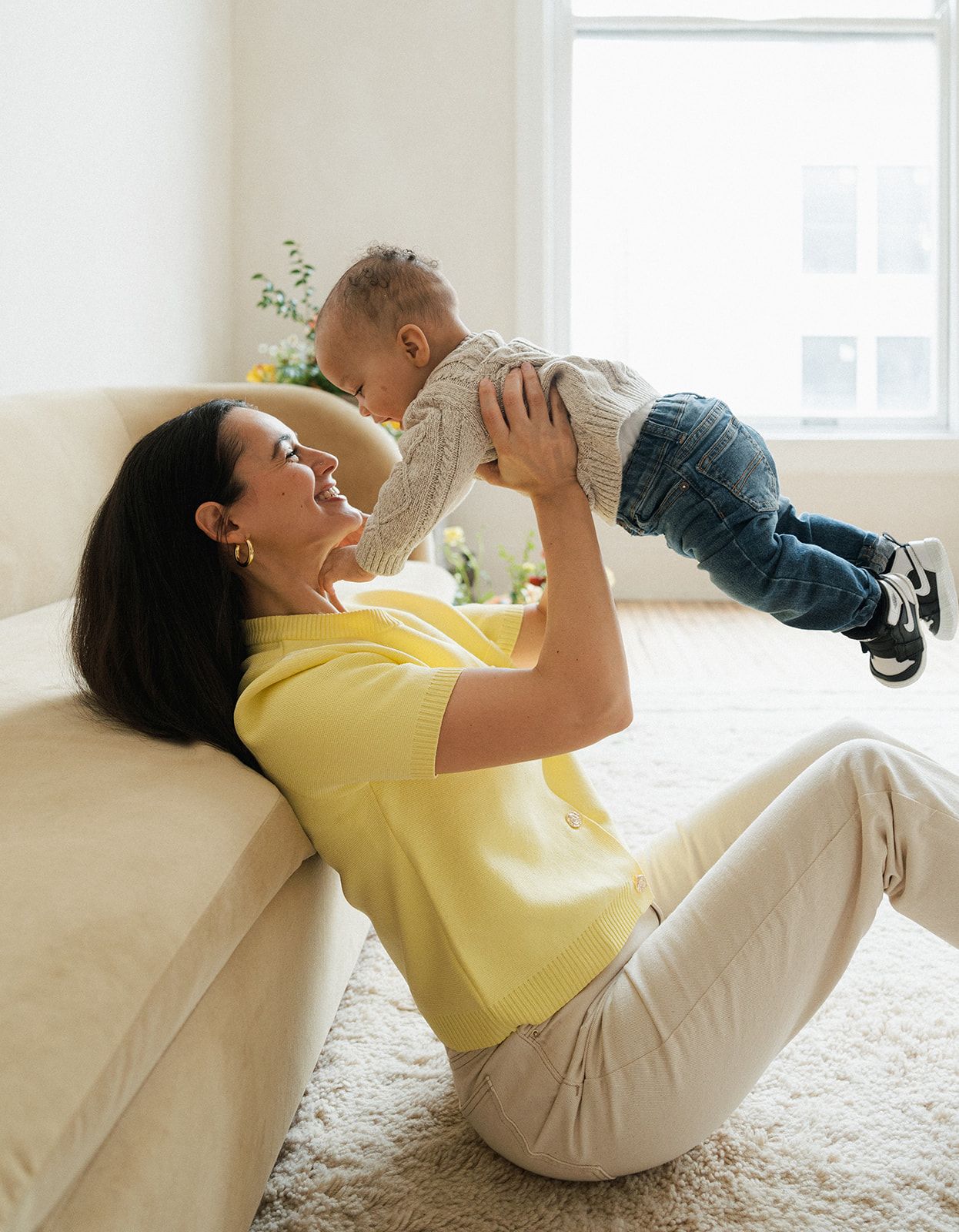 Happy mother in yellow shirt lifting smiling baby in living room, sharing joyful moment together.