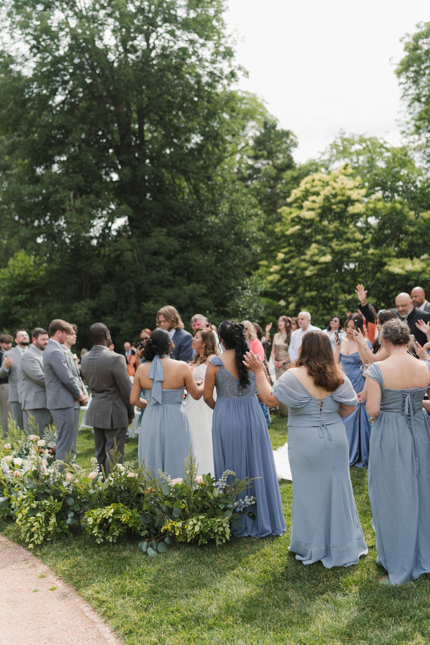 Outdoor wedding ceremony with bridesmaids in dusty blue gowns and guests applauding on a lush green lawn.