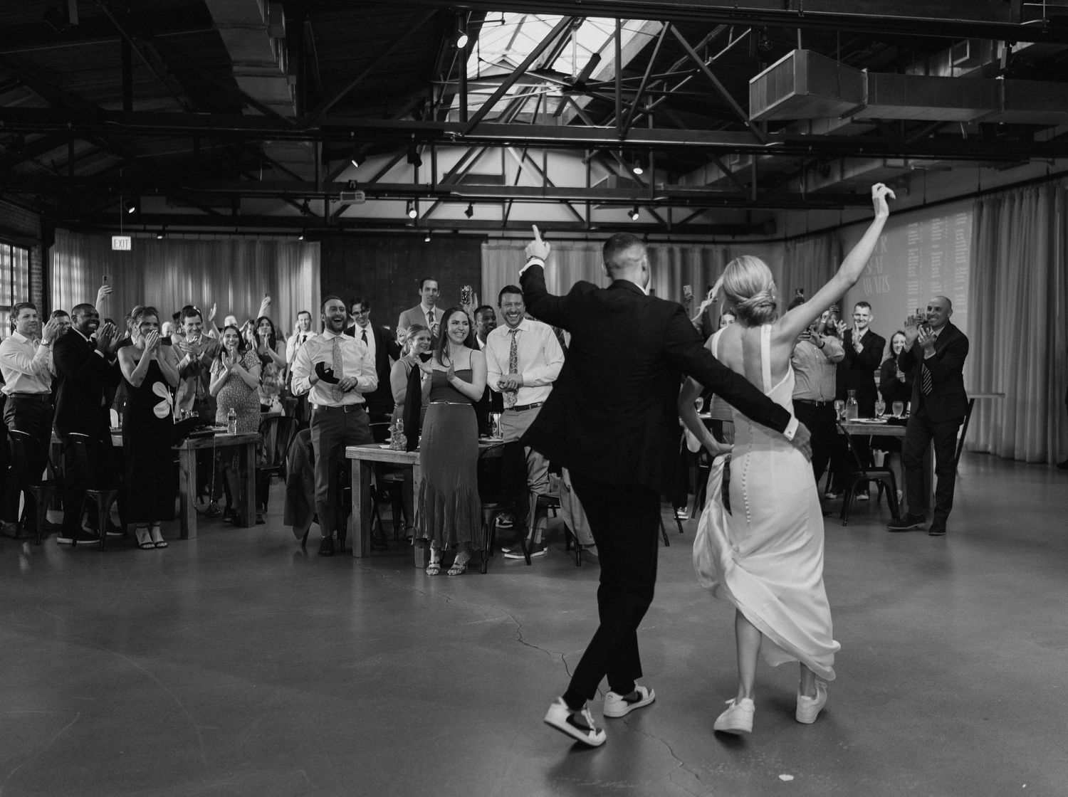 Couple shares first dance at wedding reception in industrial venue, guests applauding in black and white photo.