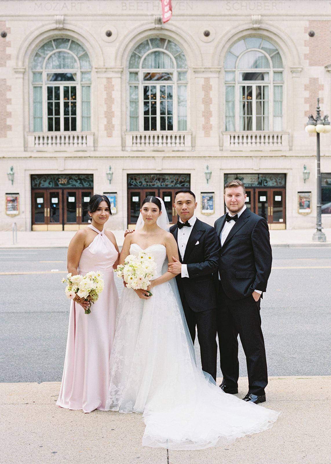 Bride and groom pose with bridesmaid and groomsman in formal attire outside an elegant neoclassical building.