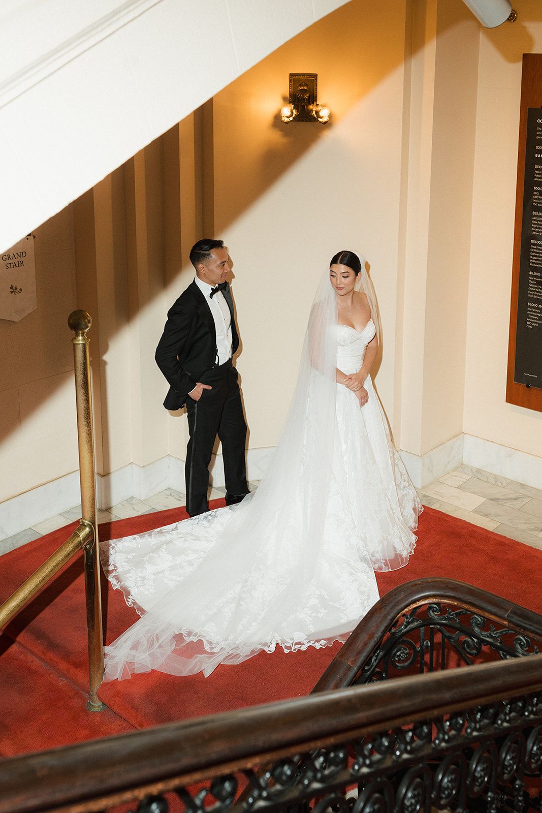 Bride and groom pose elegantly on red-carpeted staircase in classic wedding attire at formal venue.