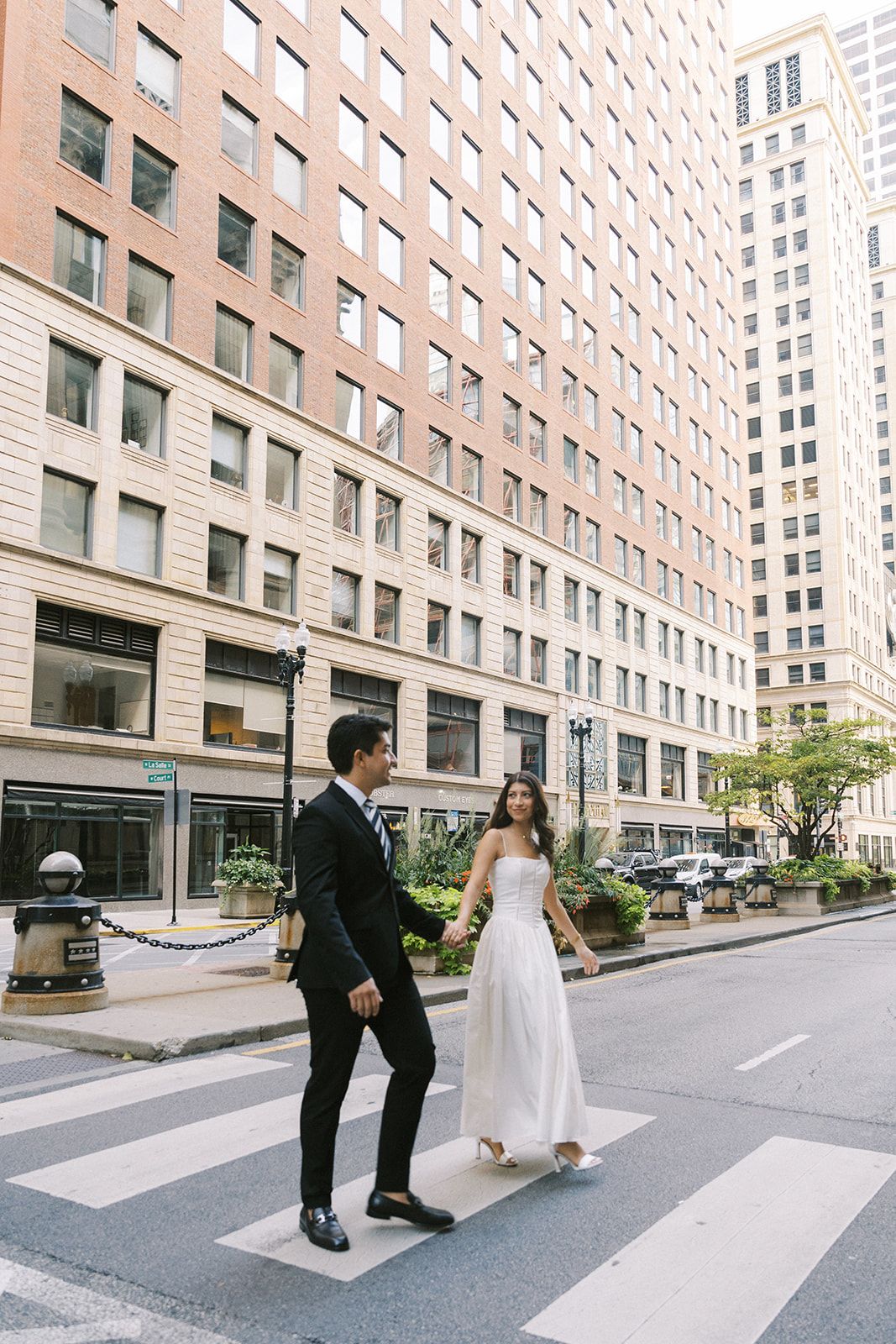 Bride and groom cross a downtown city street holding hands in wedding attire among tall buildings.