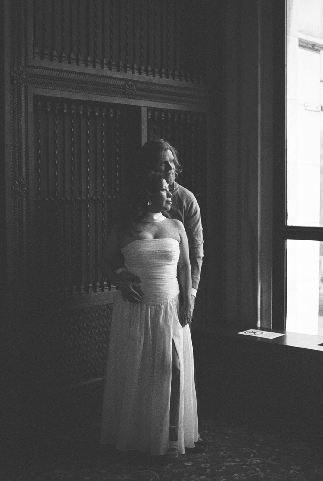 Black and white photo of a couple embracing near a window in an elegant interior, woman in a white strapless gown.