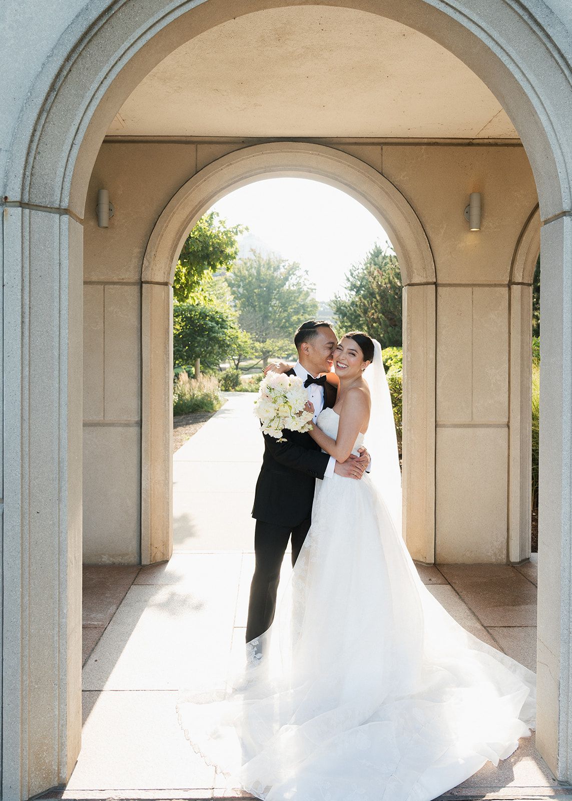 Bride and groom sharing a romantic embrace under an elegant stone archway with garden views behind them.