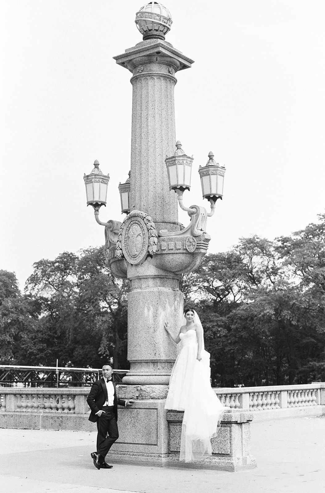 Black and white photo of bride and groom posing by ornate stone lamp post in a park.