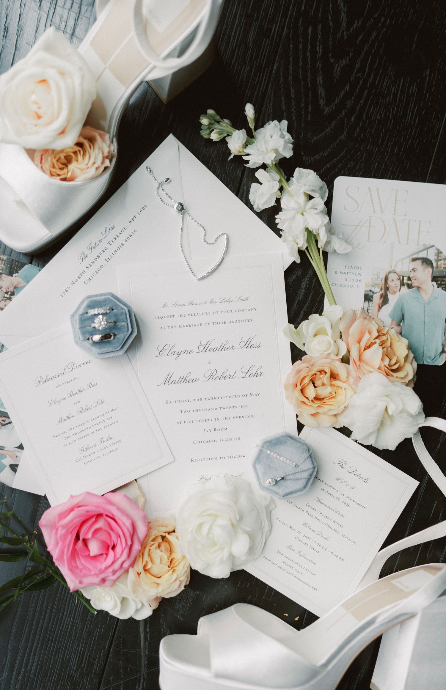 Elegant wedding invitation suite with floral arrangements, blue velvet ring boxes, white heels, and a save the date card on dark wood.