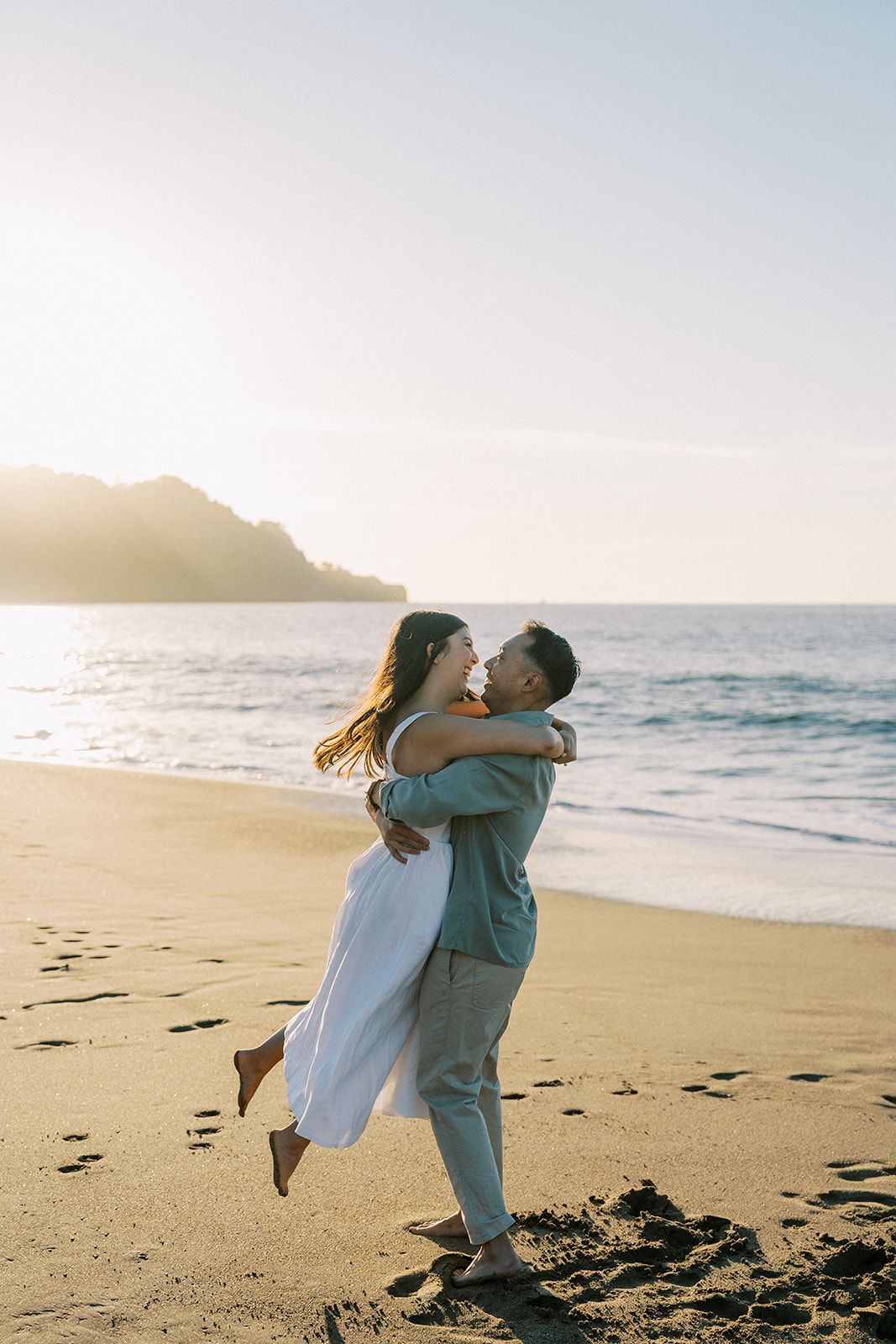 Couple embracing on sandy beach at golden sunset with ocean and mountains in background.