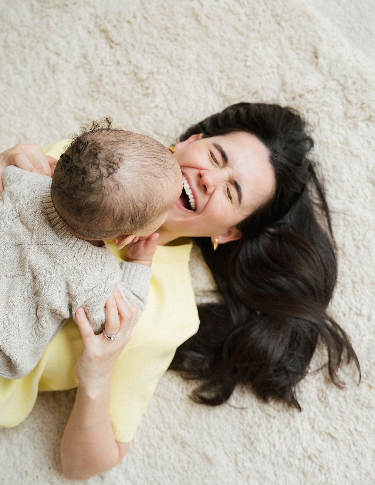 Happy dark-haired mother laughing while lying on carpet cuddling her baby in cozy sweater.