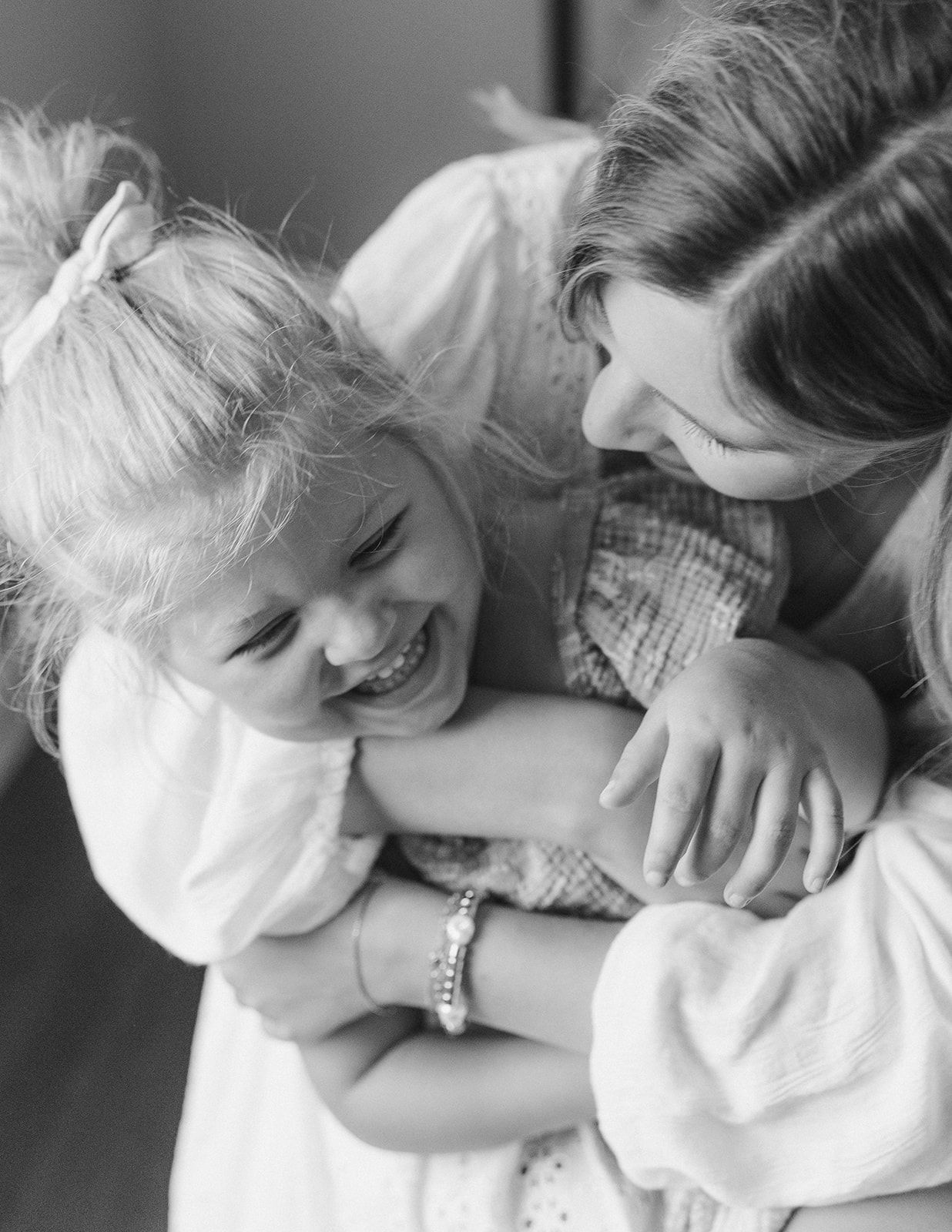 Black and white photo of a mother hugging and laughing with her young blonde daughter in white dresses.