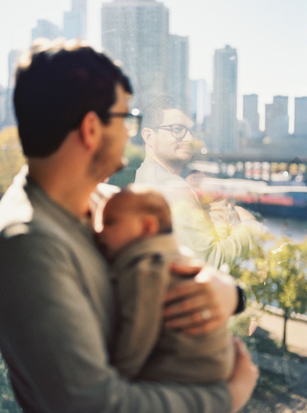 A man holding a newborn baby with a city skyline in the background on a sunny day.