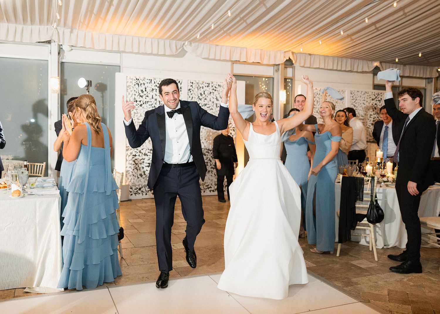 Bride and groom make grand entrance into wedding reception as guests cheer in a tented venue.