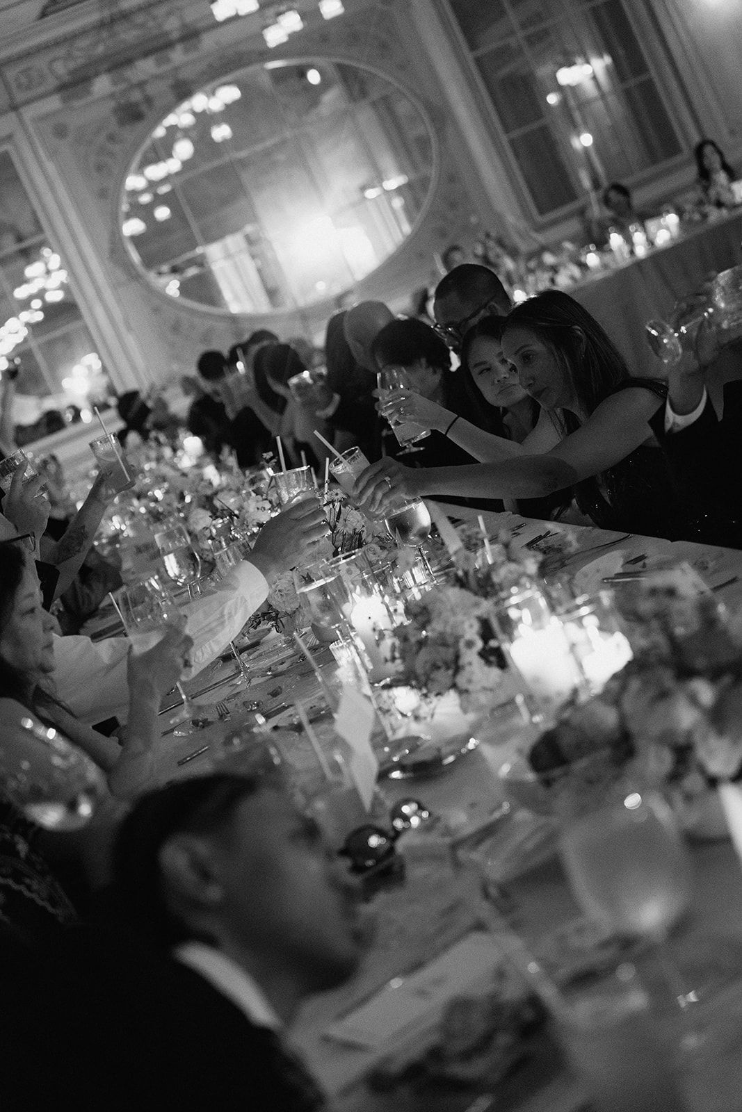 Black and white photo of elegant wedding reception guests toasting with champagne in ornate ballroom.