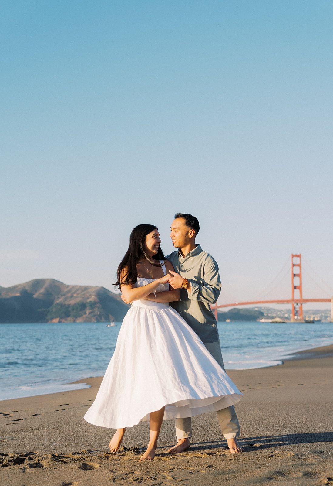 Couple dancing barefoot on Baker Beach with the Golden Gate Bridge in the background at sunset.