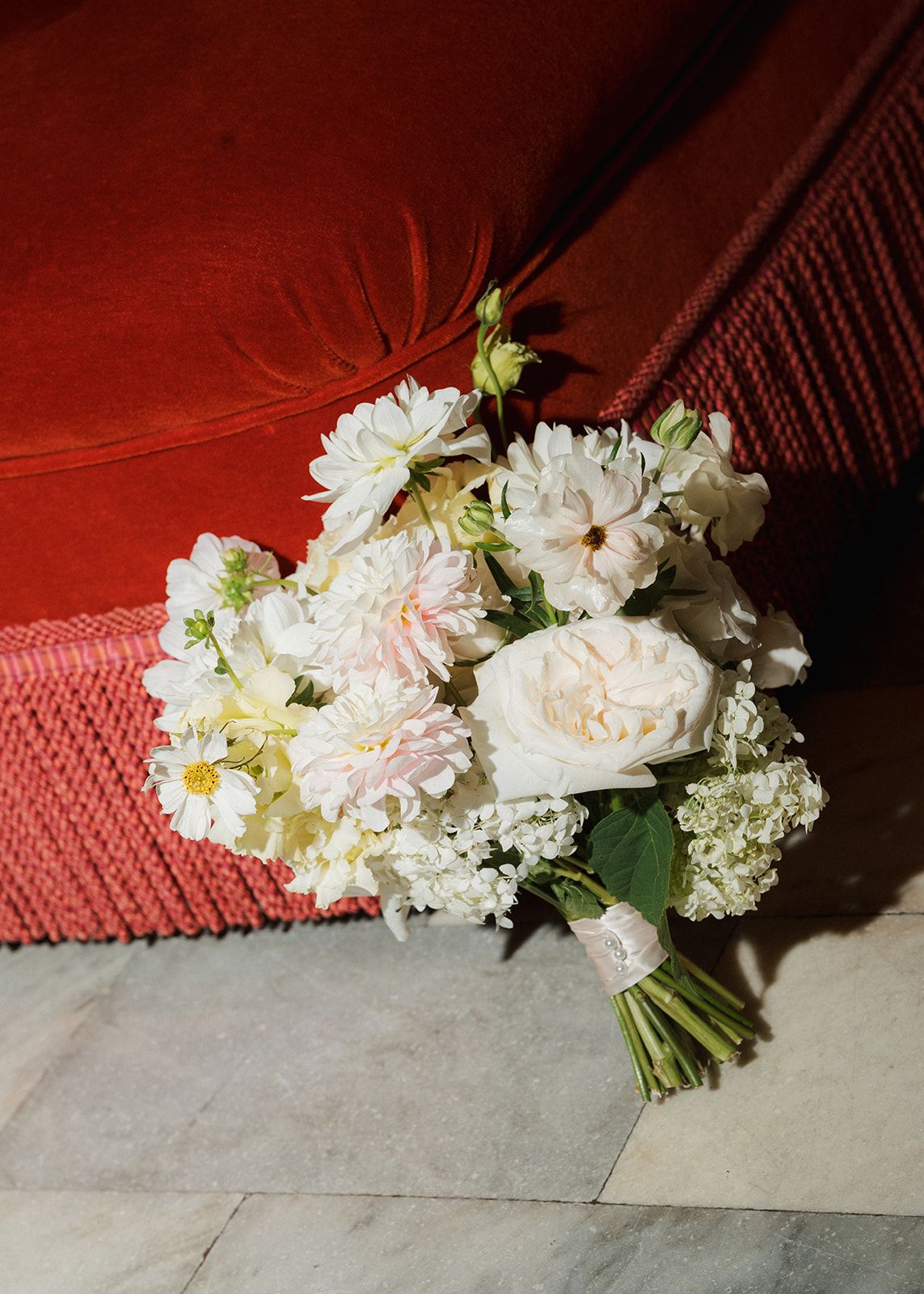 Romantic bridal bouquet of white roses, daisies, and peonies rests against a red velvet sofa.