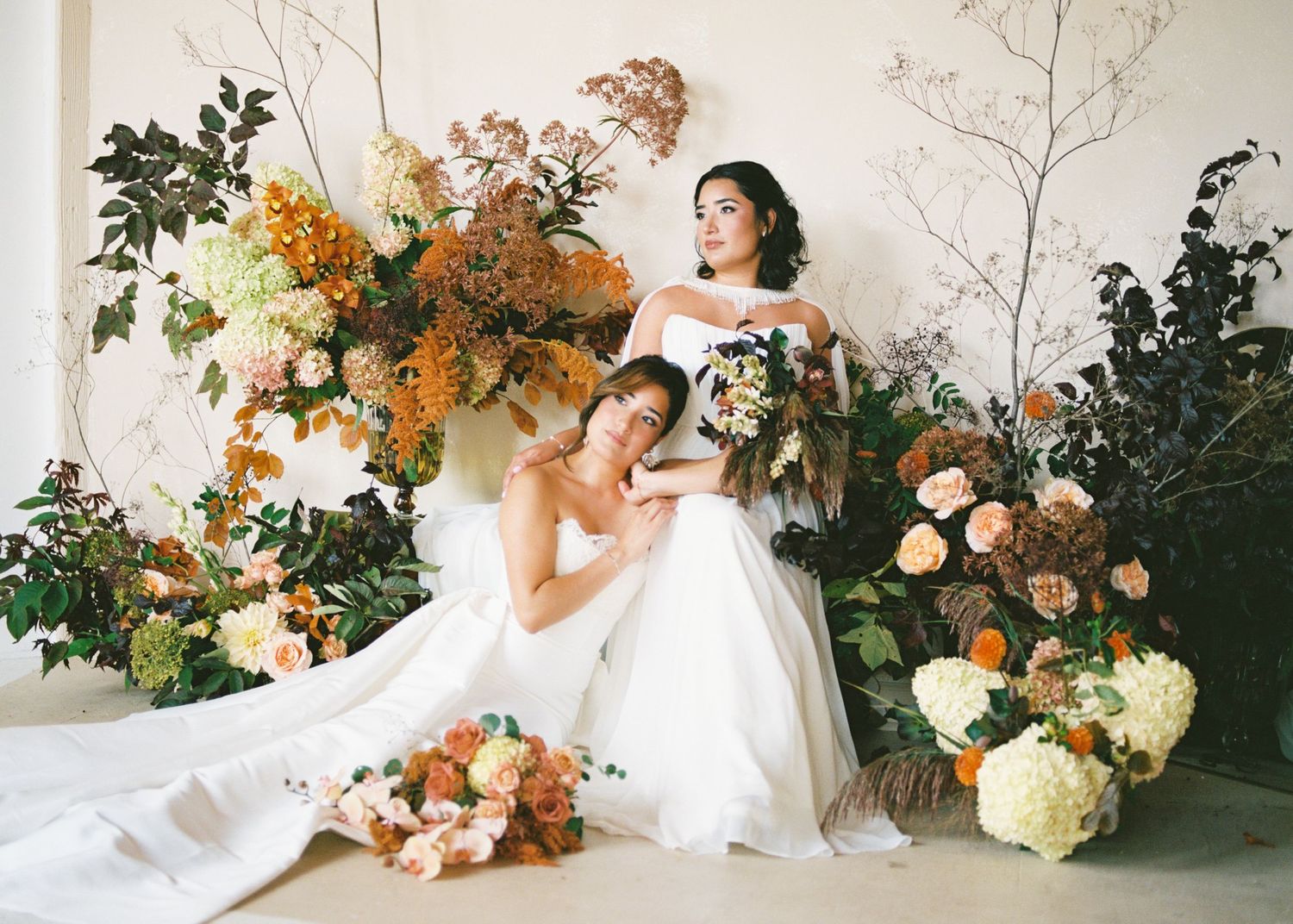 Two brides in white gowns pose among lush autumn floral arrangements of orange, rust, and cream blooms.