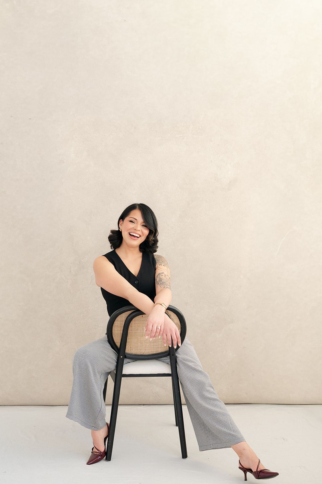 Smiling woman with tattoos sits on a cane chair wearing black top and plaid trousers against beige wall.