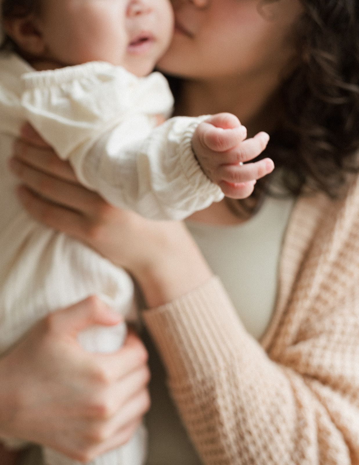 Mother in beige knit sweater kissing her newborn baby while holding infant up close.