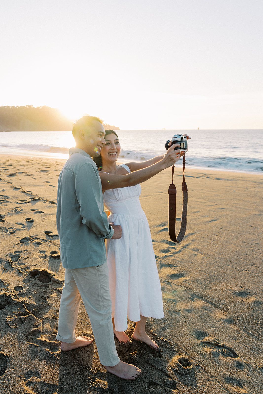 Couple taking a selfie with film camera on a sandy beach at golden sunset hour.
