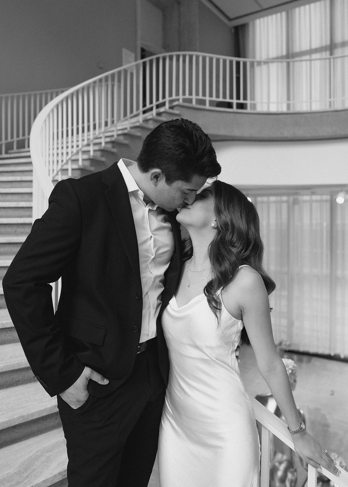 Black and white photo of a couple in wedding attire sharing a kiss near an elegant staircase railing.