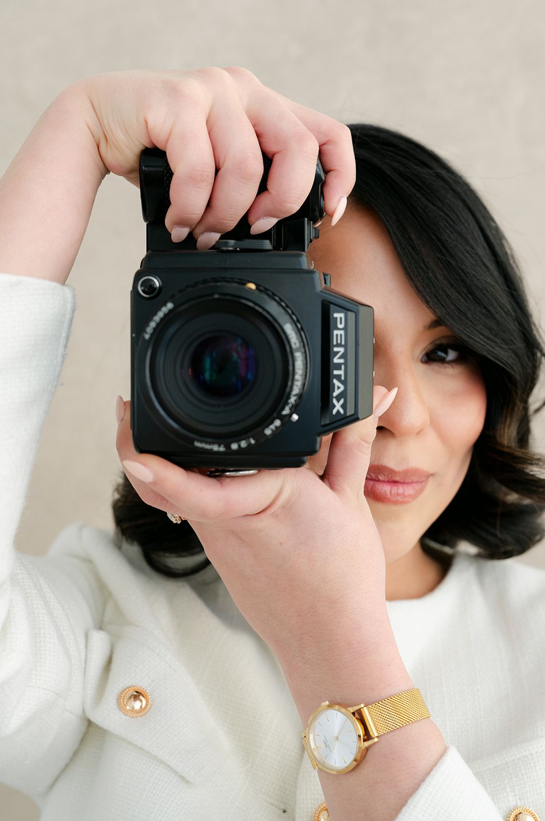 Woman in white jacket holding a black Pentax medium format camera up to her face while wearing a gold watch.