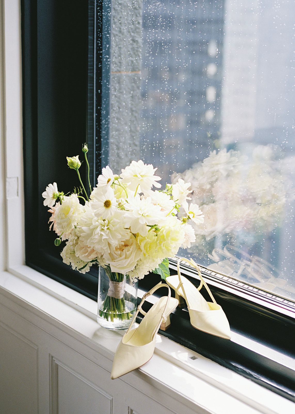 White wedding bouquet in glass vase beside ivory heeled shoes on windowsill with rain-speckled glass.