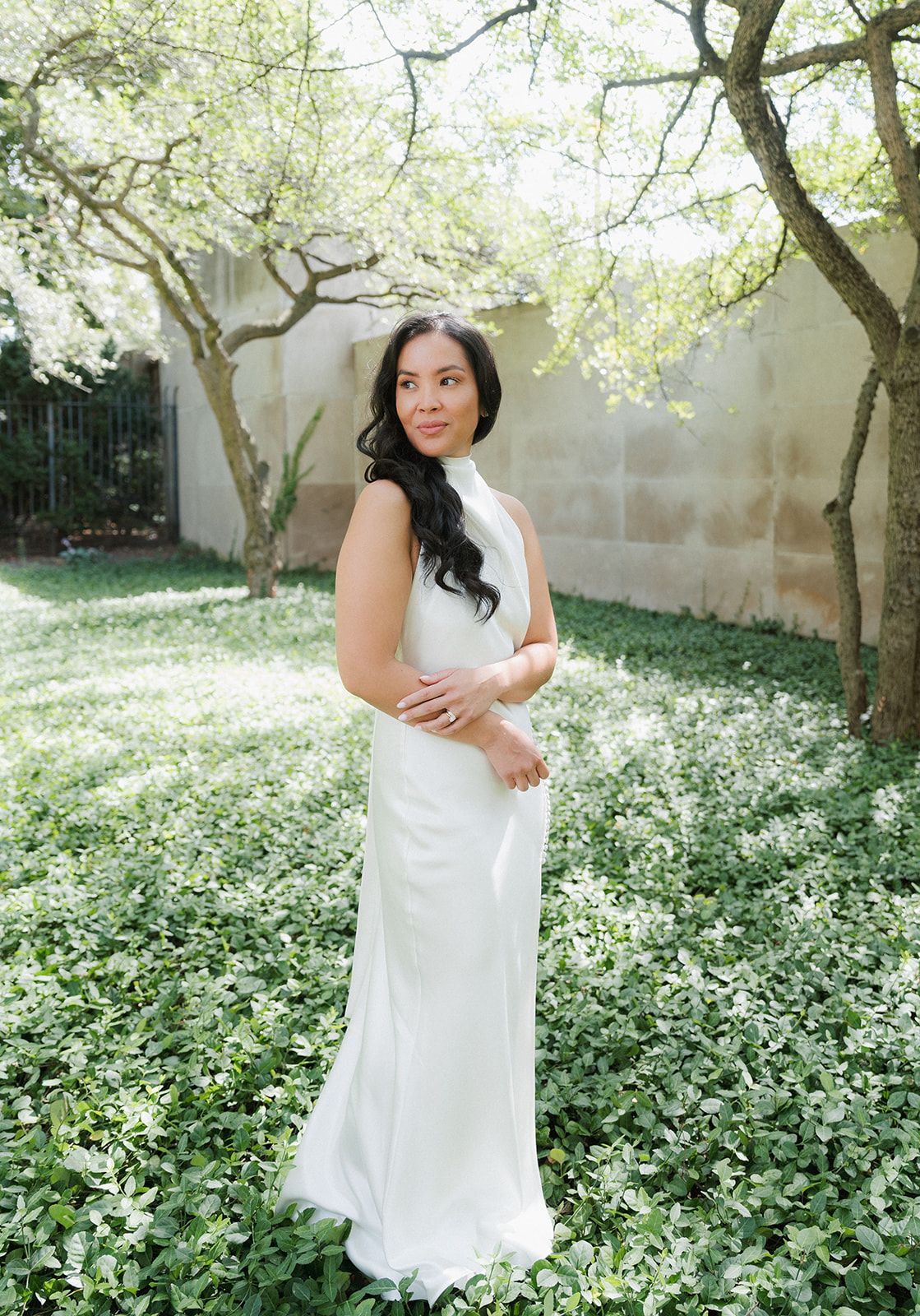 A bride in a sleek white halter wedding gown poses gracefully among lush green ground cover outdoors.