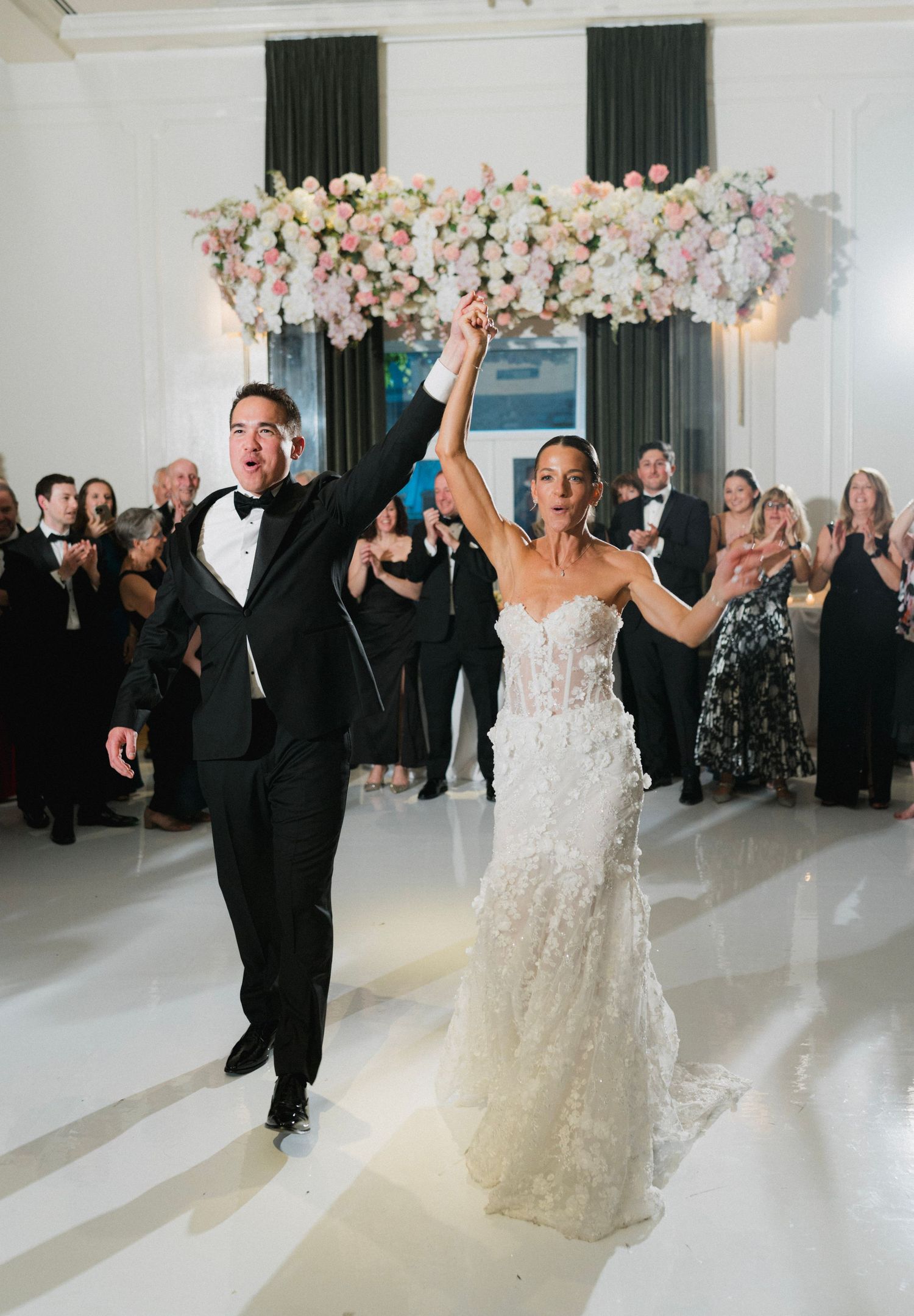 Bride and groom raise hands triumphantly at wedding reception with floral backdrop and cheering guests.