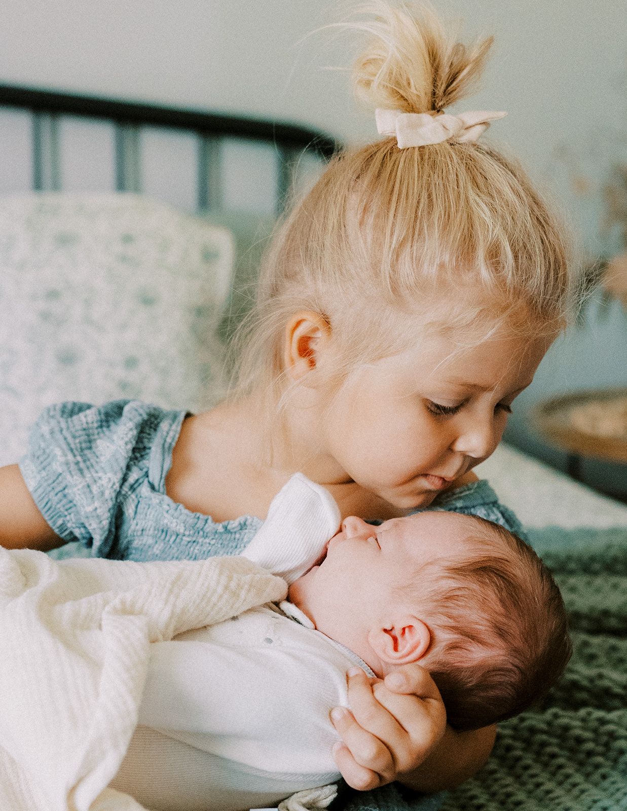 Young blonde girl with hair bow gently kissing sleeping newborn baby held in her arms.