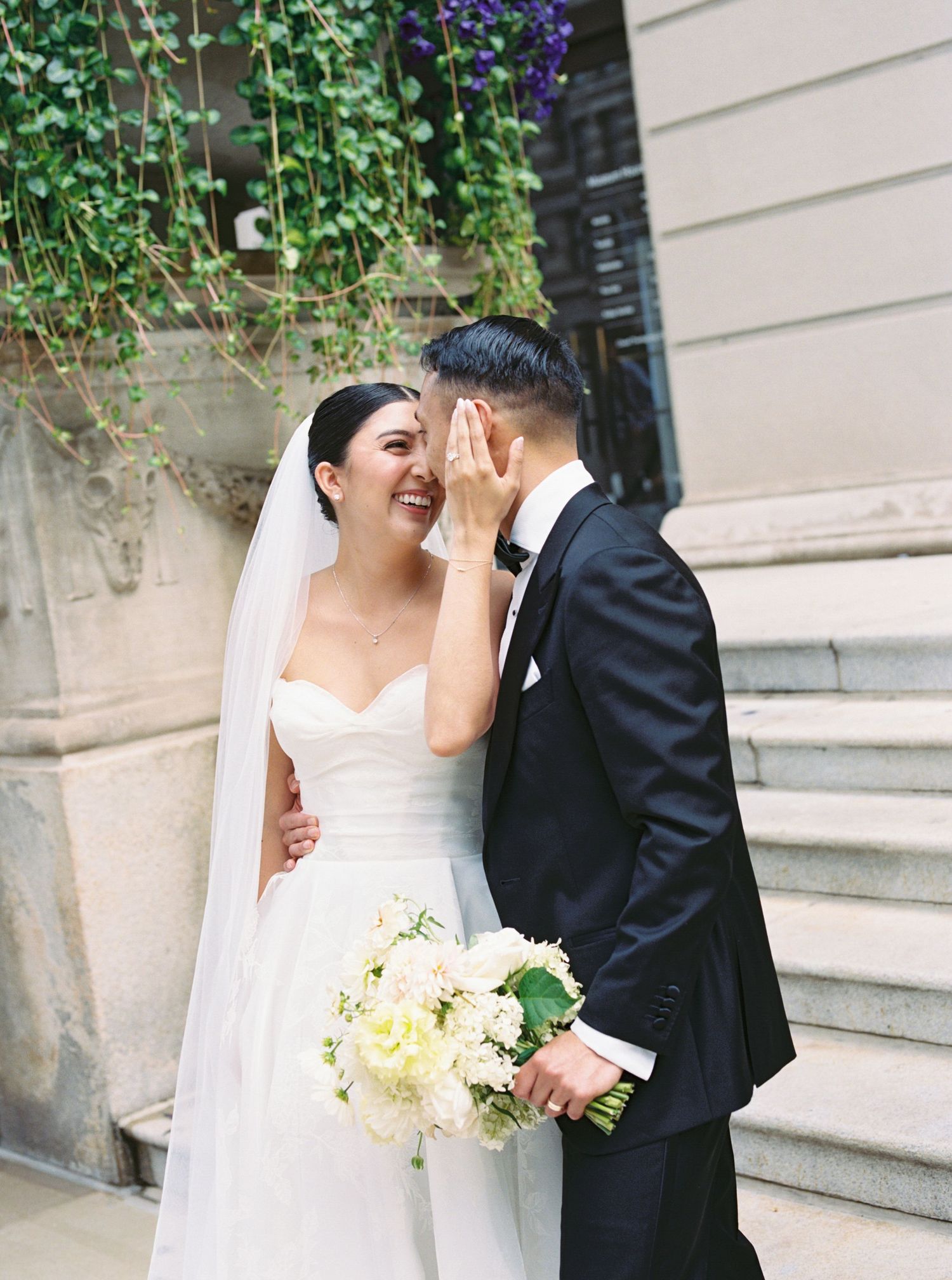 Laughing bride in strapless white gown with veil holds white bouquet as groom whispers to her on stone steps.