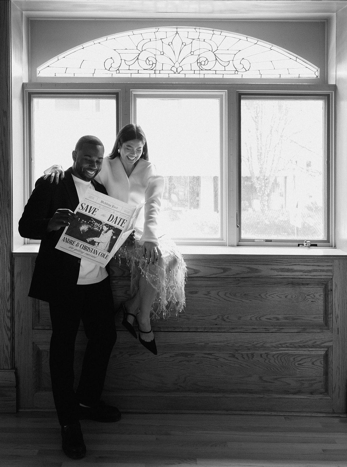 Black and white photo of smiling couple holding Save the Date newspaper announcement near large window.