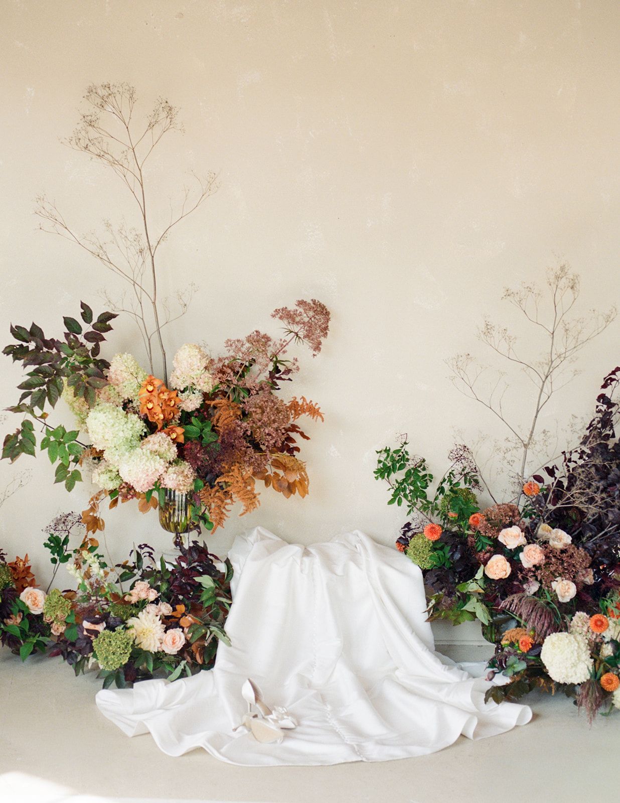 Artistic autumn wedding floral installation with rust, peach, and burgundy blooms against neutral wall.