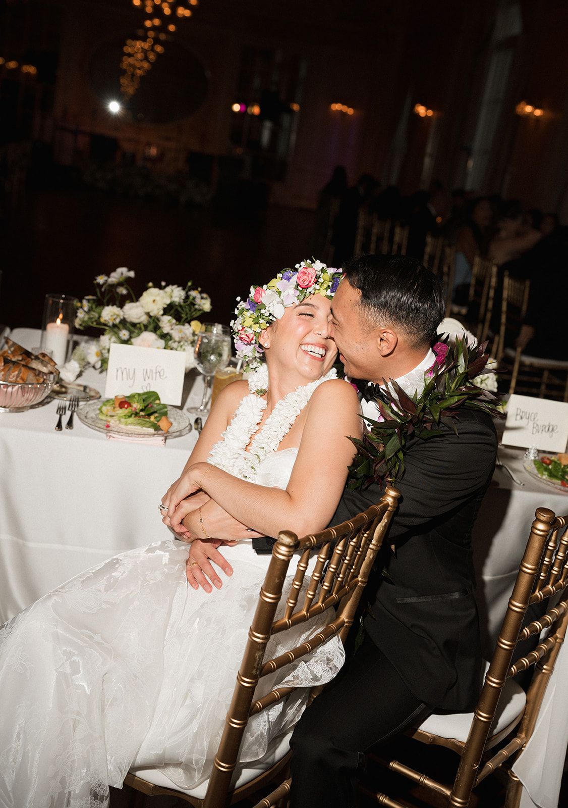 Joyful bride wearing floral crown laughs as groom kisses her cheek at wedding reception sweetheart table.