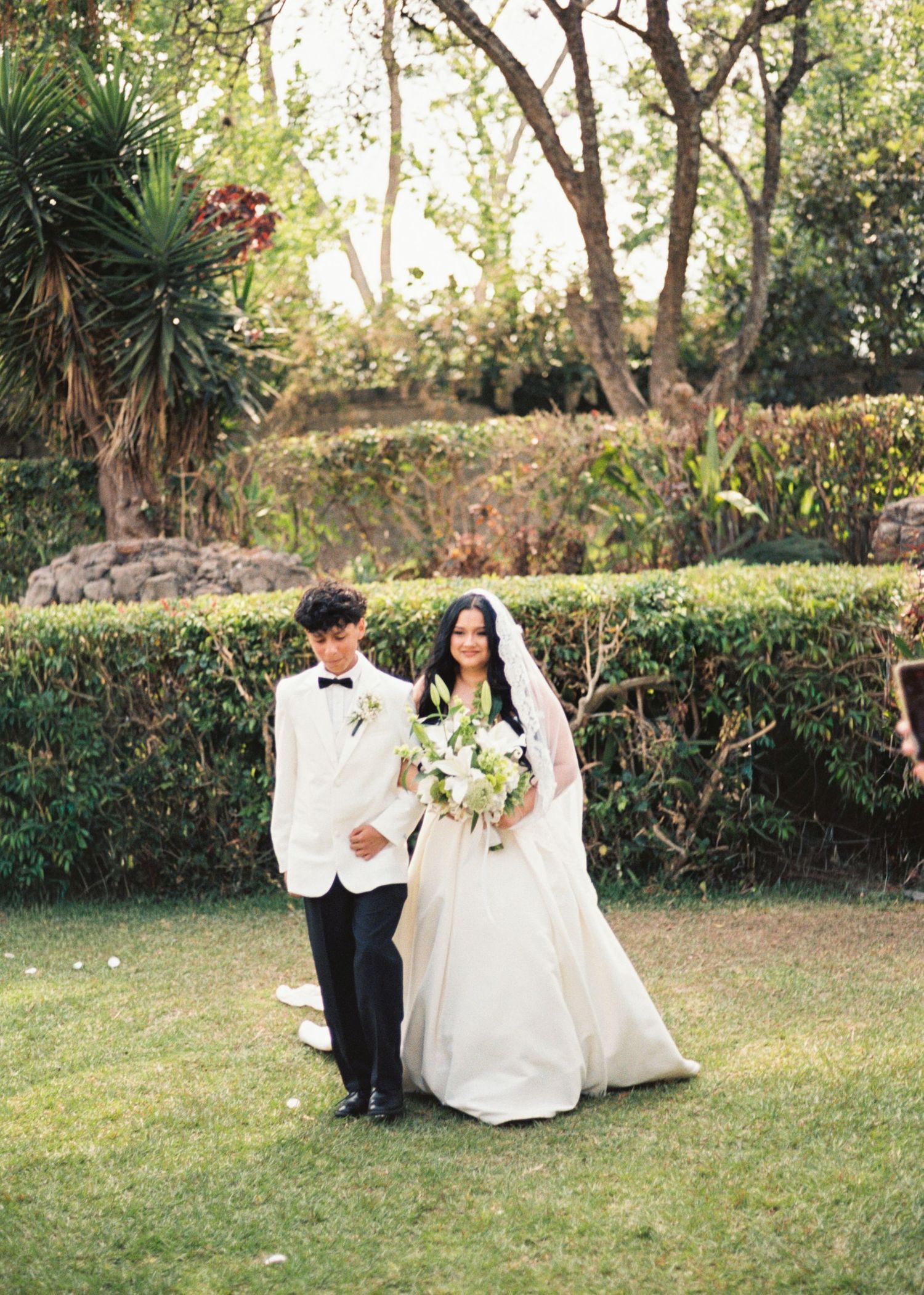 Bride in white gown holding bouquet stands with groom in white tuxedo jacket on lush garden lawn.