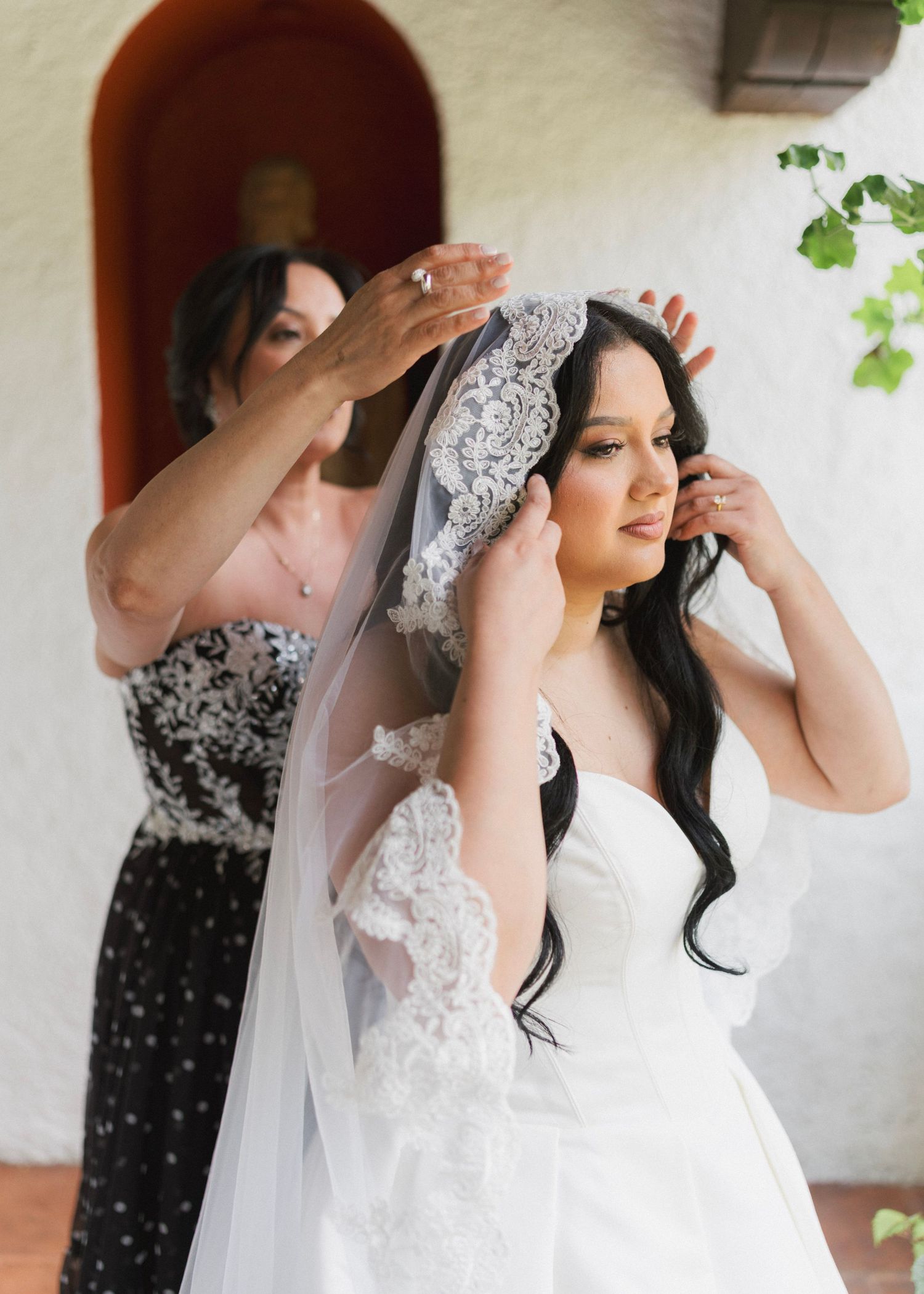 A bride in a white gown gets her lace veil placed by a woman in a black dress before her wedding ceremony.