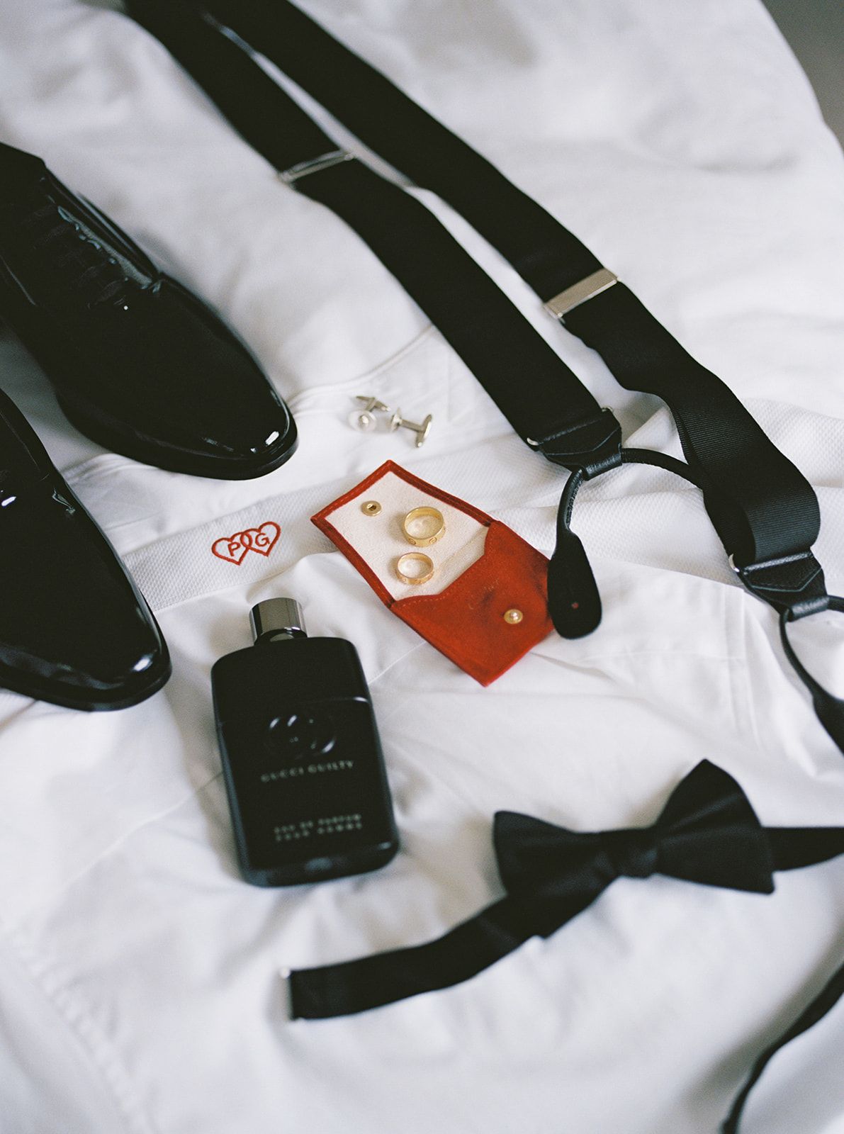 Groom wedding flat lay featuring black patent shoes, suspenders, bow tie, cologne, cufflinks, and gold rings.